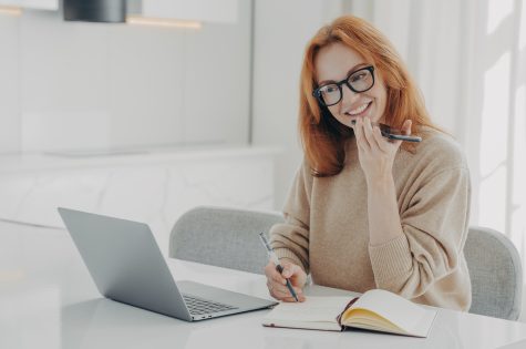Positive ginger woman in casual clothes records audio message or smartphone, talks with virtual digital voice assistant while working on laptop, sits at her workplace, female uses speakerphone at work